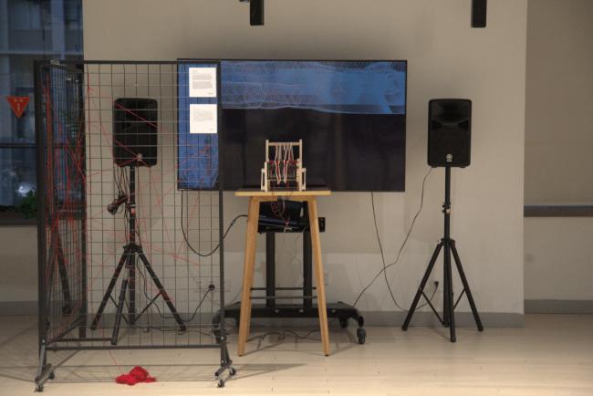 The set up includes a TV screen, two speakers, a table, a weaving loom with sensors and switches and threads, and a pair of standing grids with tangled threads.